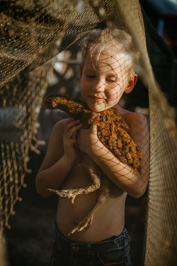 Shirtless boy holding chicken while standing outdoors