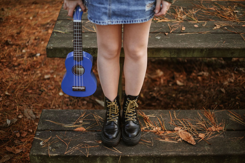 High angle of crop teenager in blue denim skirt and black leather boots standing alone on old wooden bench beside table among brown dry leaves and pine needles and holding small blue guitar in autumn forest