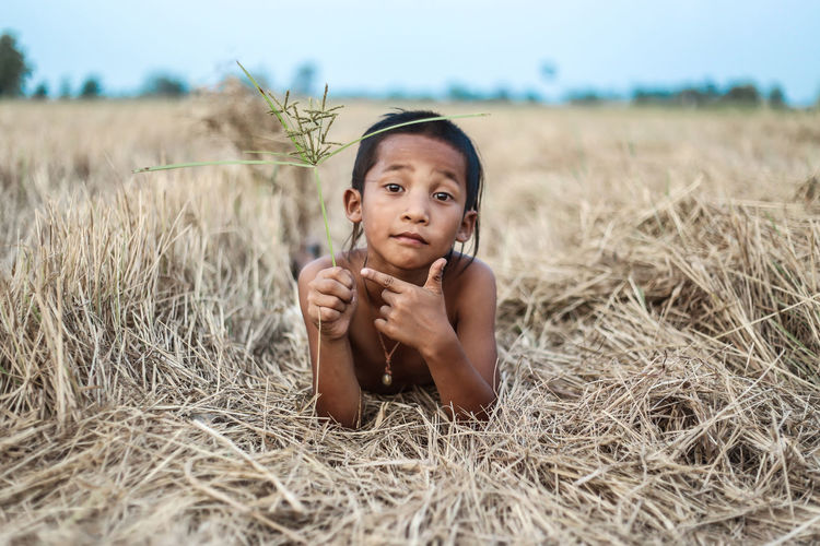 Portrait of boy holding grass gesturing while lying on field against sky at farm
