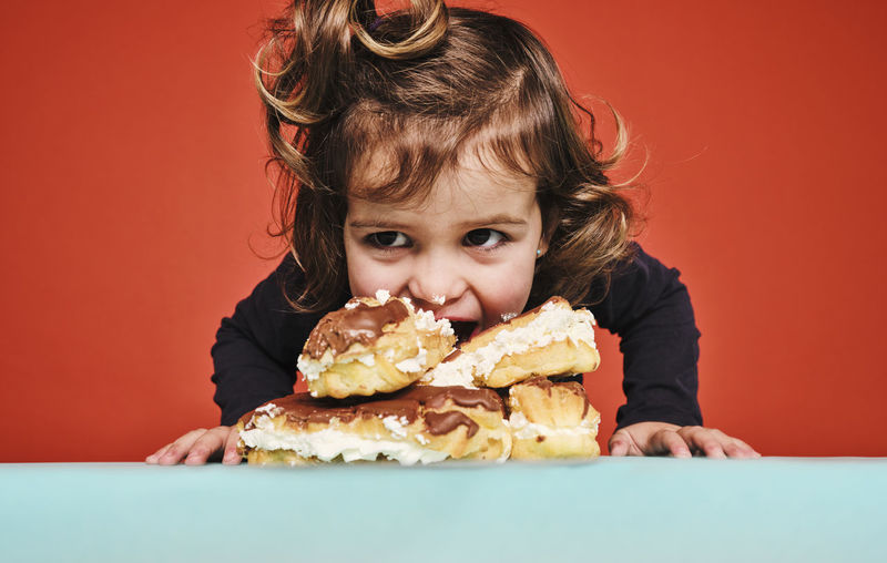 Closeup portrait of cheerful little girl enjoying sweet eclairs with chocolate while looking away sitting on the table against red background