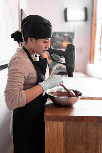 Side view of focus ethnic female worker in black apron and cap wearing gloves while melting dark chocolate in stainless steel bowl using heat gun working at counter with marble slab