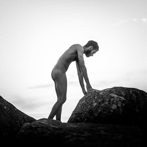 Side view of shirtless man standing on rock against sky