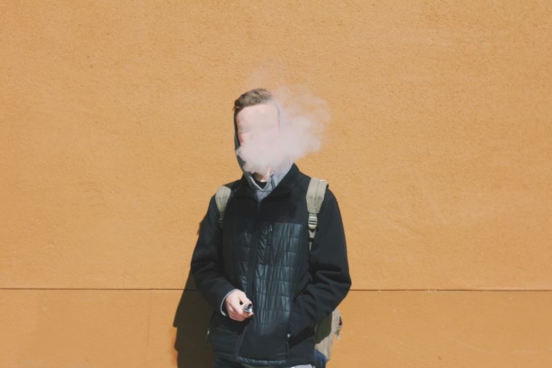 Young man smoking while standing against wall on sunny day