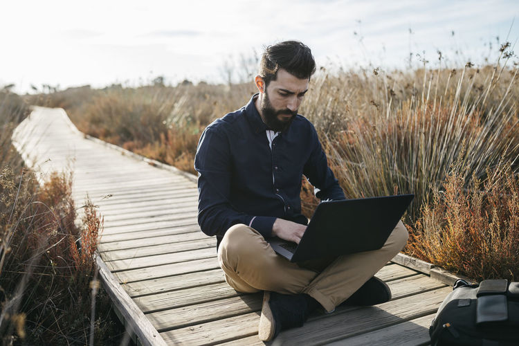 Man using laptop while sitting on boardwalk against sky