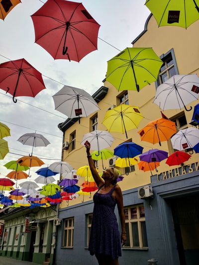 Woman reaching multi colored umbrellas while standing against buildings