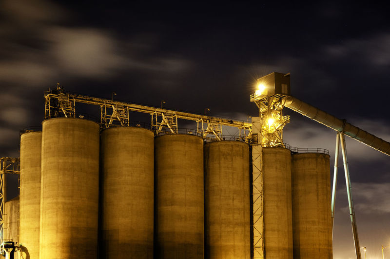Low angle view of illuminated storage tanks at industry against cloudy sky