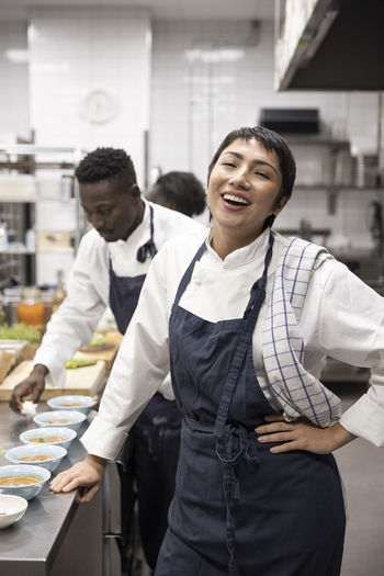 Portrait of chef laughing while colleague wiping bowls in restaurant kitchen
