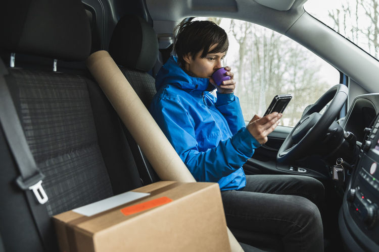 Female essential service driver drinking coffee while using smart phone in delivery van