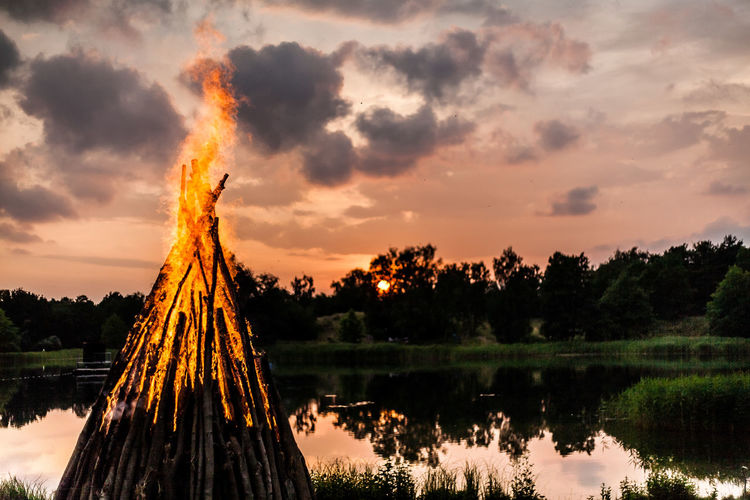 Bonfire by pond against cloudy sky during sunset