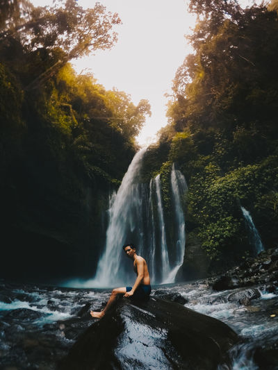 Side view of shirtless man sitting against waterfall in forest