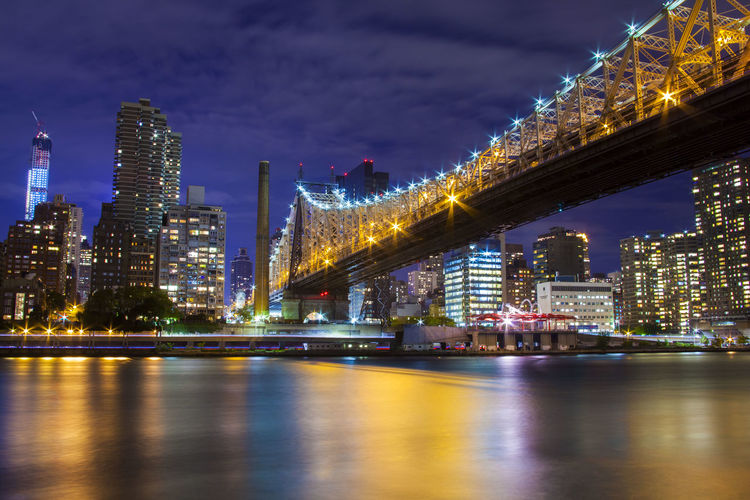Low angle view of illuminated queensboro bridge over river against buildings at night