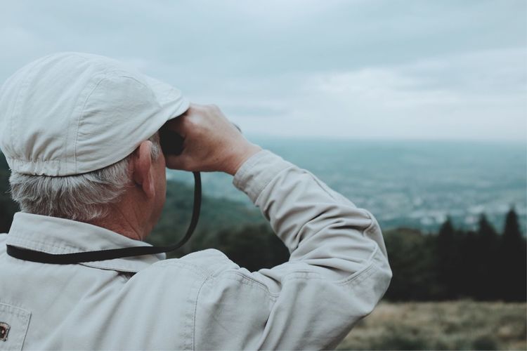 Rear view of senior man looking through binoculars while standing on mountain against cloudy sky