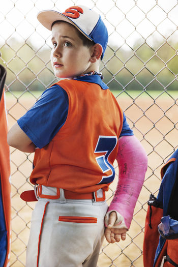 Side view of baseball player with fractured hand standing by chainklink fence on field