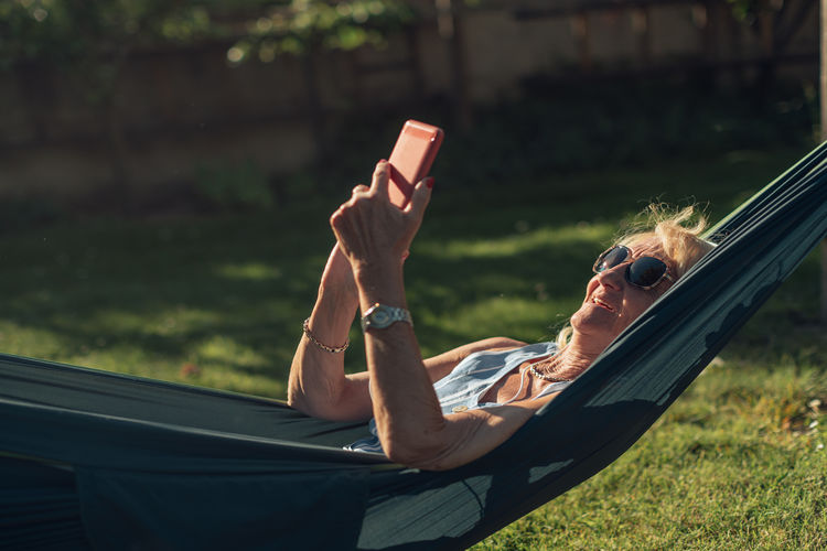 Caucasian senior lady swings in the hammock listening to music with phone and headphones.