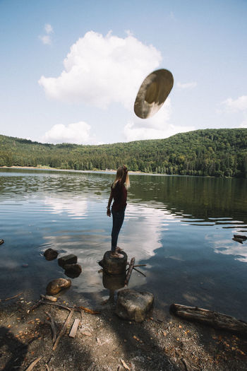 Full length of young woman throwing hat while standing by lake
