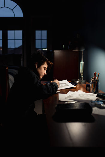 Teenage boy drawing at a desk in a dark room by lamp light.