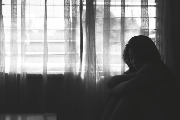 Sad young woman hugging knees against curtains at home