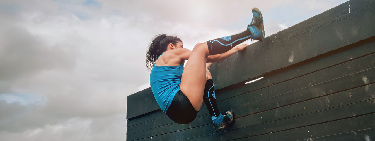 Low angle view of sporty woman climbing wooden wall against sky