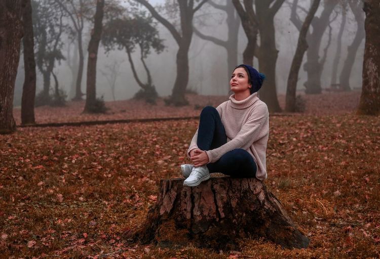 Full length of thoughtful young woman sitting on tree stump in forest during autumn