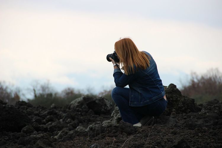 Rear view of woman photographing through camera while crouching on field