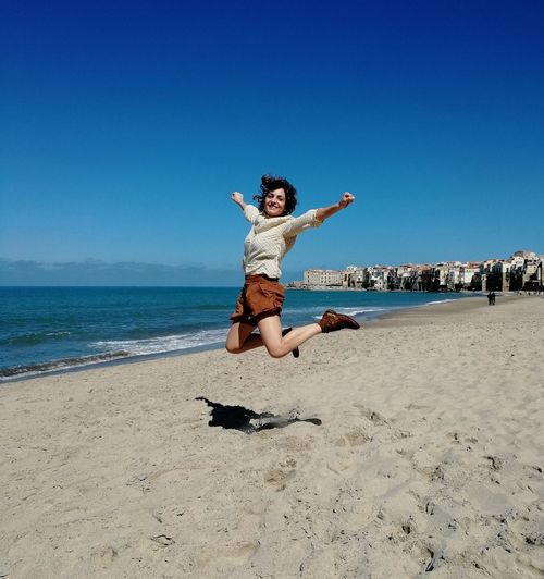 Full length of woman jumping at beach against clear blue sky