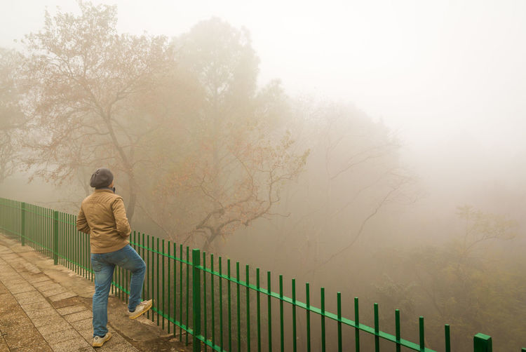 Rear view of man wearing warm clothing standing by railing during foggy weather