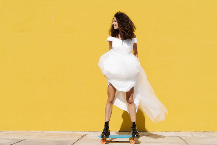 Full body of happy cheerful bride in white dress and boots standing on longboard against yellow background in sunlight