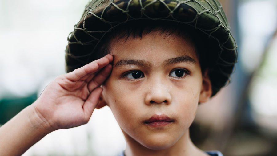 Close-up of cute boy wearing helmet looking away while saluting outdoors