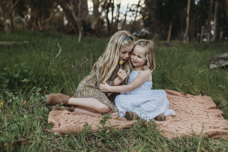 Two blond sisters sitting on a blanket in a field with lollipops