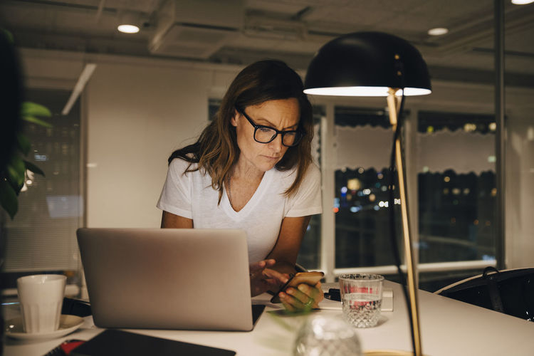 Confident female entrepreneur using smart phone while sitting with laptop at illuminated desk in workplace during night