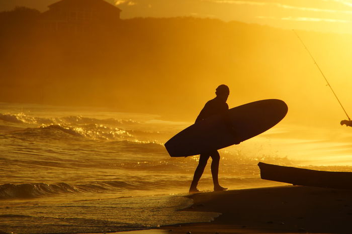 Silhouette man with surfboard at sea shore during sunset