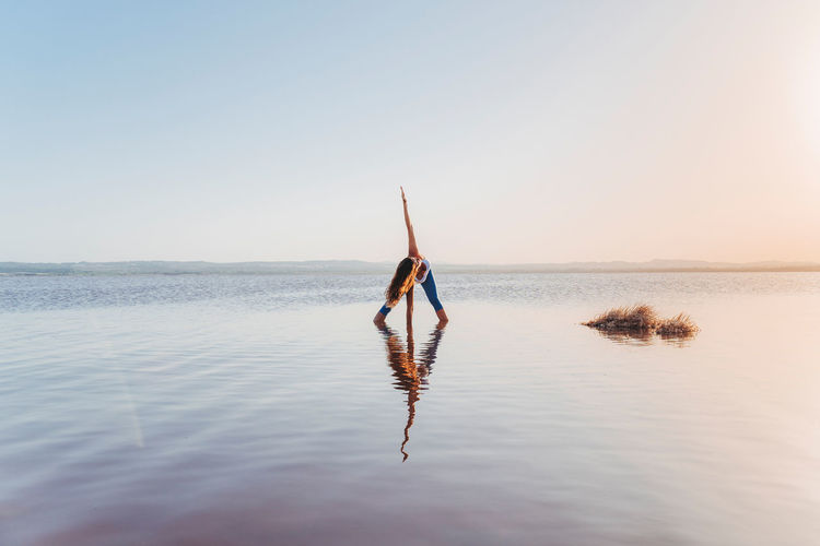 Full body of unrecognizable flexible female in sportswear doing revolved wide legged forward bend while standing in calm lake water and practicing yoga in summer evening