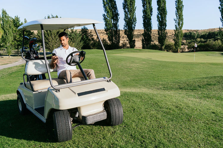 Adult male athlete text messaging on cellphone in vehicle with golf equipment on course in sunlight