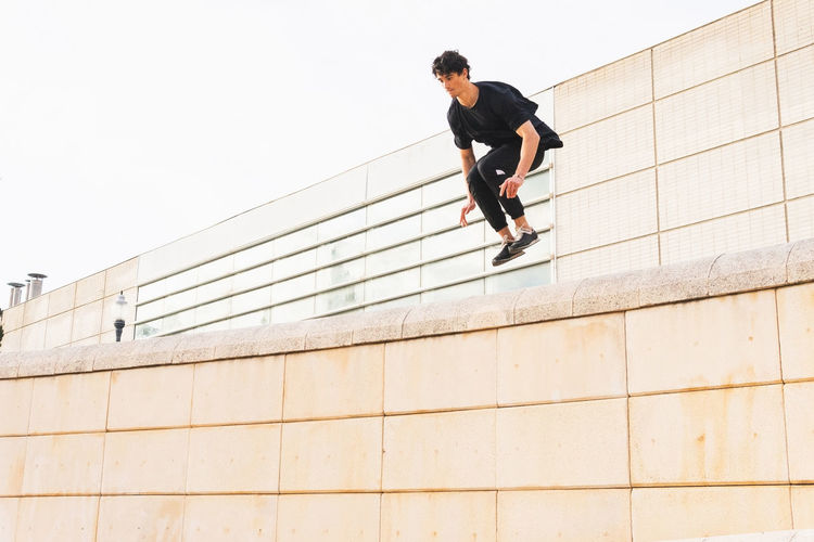 Low angle of full length athlete man in sports outfit doing jump above wall and training parkour stunt