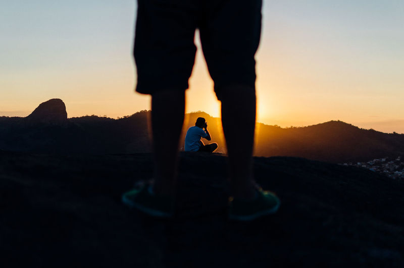 Low section of man standing on rock against photographer during sunset