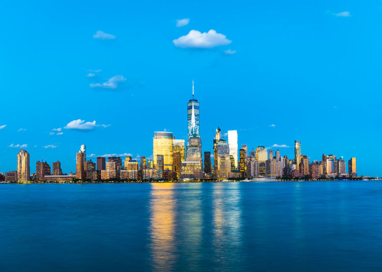 Illuminated one world trade center and buildings in city by hudson river against blue sky