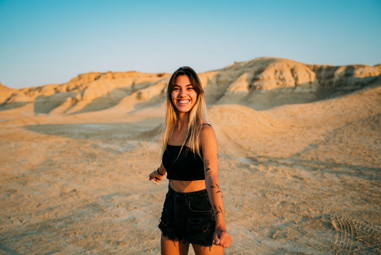 Positive female traveler standing in dry valley in bardenas reales and enjoying sunset in summer while looking at camera