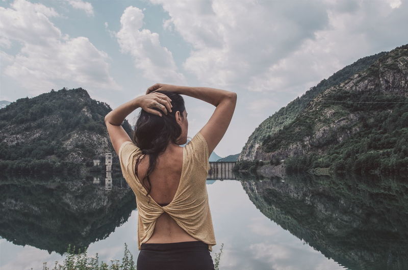 Rear view of woman with hands in hair standing by lake