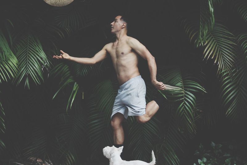 Shirtless man catching sun hat by white dog against trees
