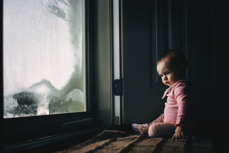 Baby girl sitting by condensed glass door at home