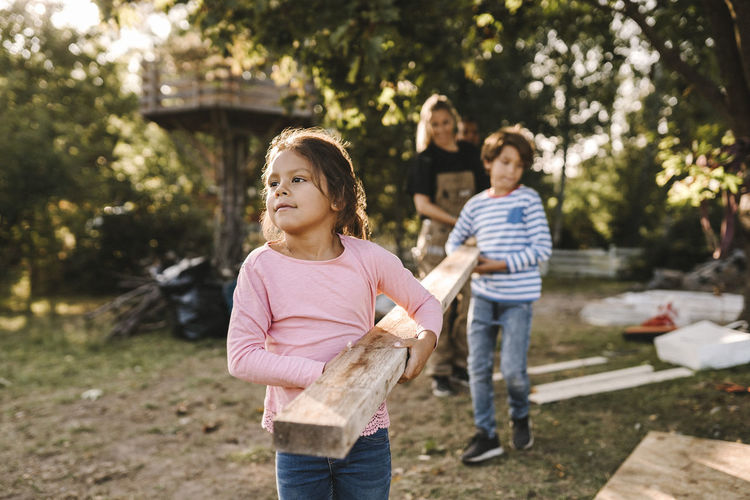 Confident girl with family carrying wooden plank in yard