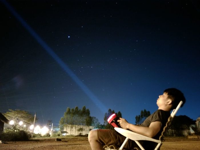 Side view of young man sitting on land against sky at night