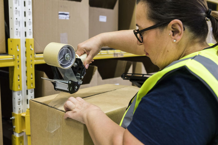 Female worker packing cardboard box with adhesive tape at distribution warehouse