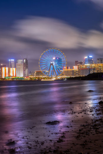 Illuminated ferris wheel by buildings against sky at night