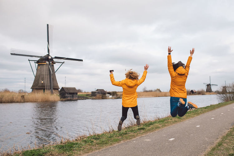 Rear view of two women jumping against sky and traditional windmills 