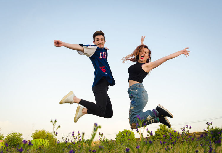 Full length side view of excited young couple jumping against sky on field