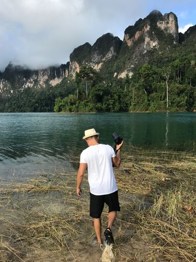 Rear view of man holding camera while walking in lake against mountains and sky