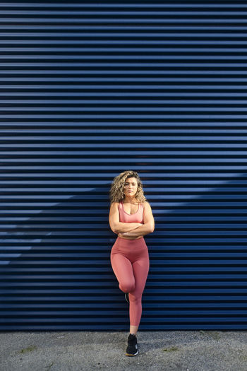 Confident sportswoman with arms crossed leaning on blue wall