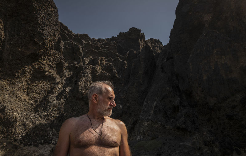 Portrait of shirtless man standing on beach against rock