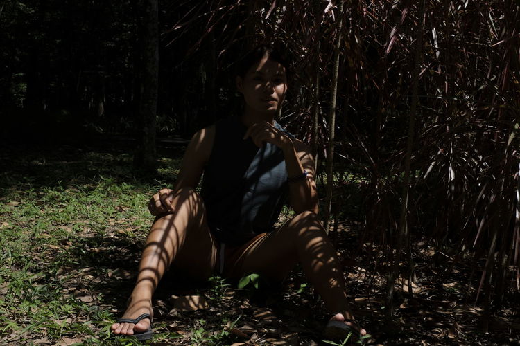Thoughtful woman sitting under trees shadow in forest
